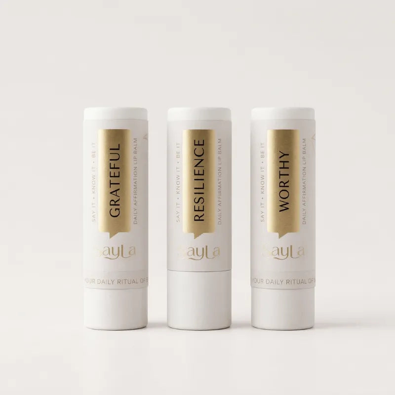 Three lip balms in 'Grateful', 'Resilience', and 'Worthy' variants with gold text on a light gray background.